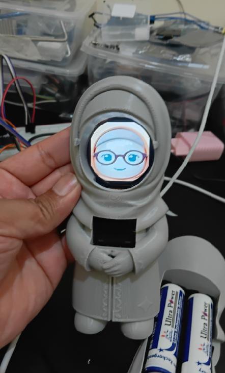 Character toy prototype held at the workbench, with an illustrated face displayed on its screen